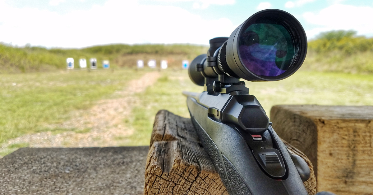 A rifle with scope resting on a wooden block at a shooting range during the summer.
