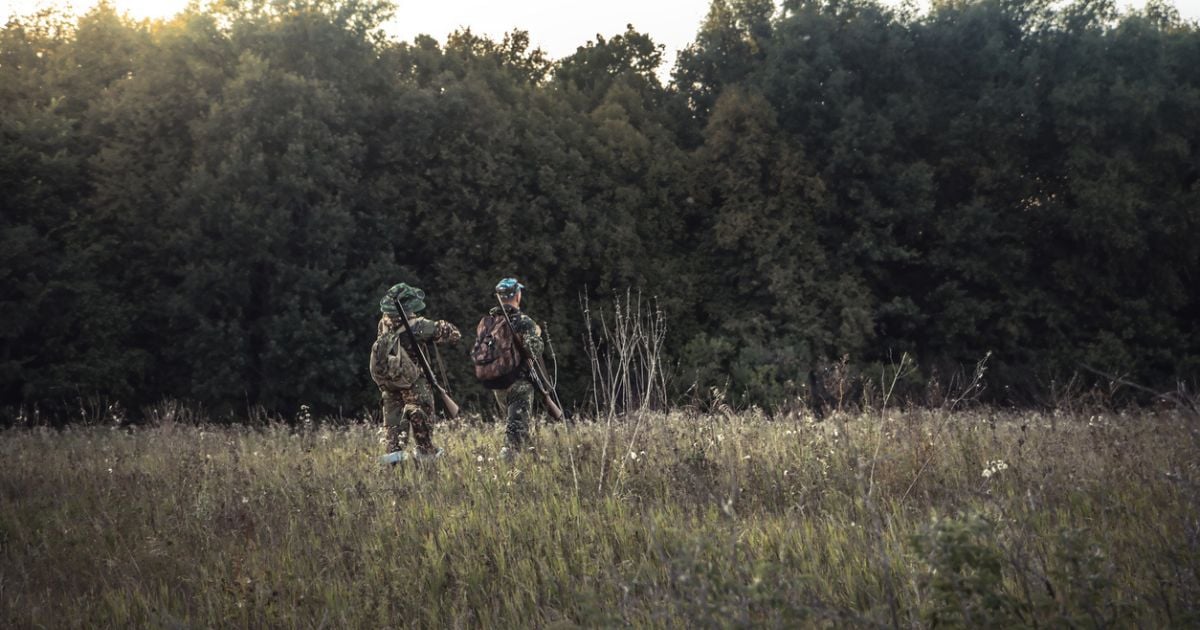 Two men hiking through grass carrying western hunting gear.