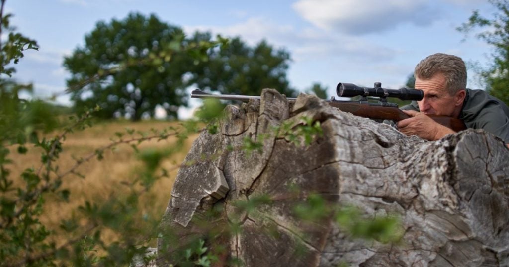 A hunter with his rifle proped on a tree trunk for stability while shooting.